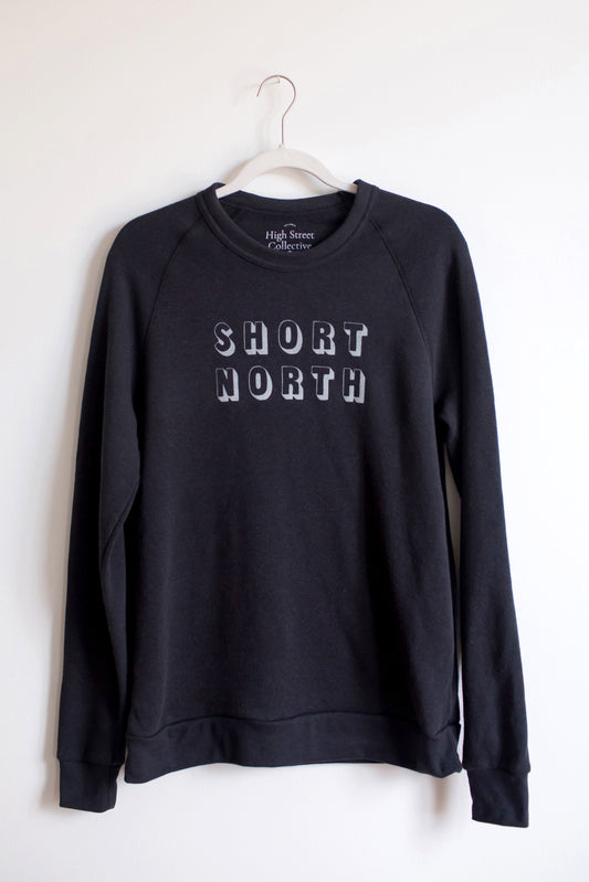 Hanging black, slim fit, crewneck sweatshirt with white Short North block graphic at front. This item has raglan sleeves for a vintage look.