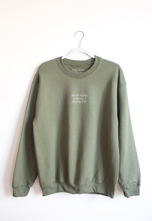 Hanging military green crewneck sweatshirt with white Short North coordinates graphic at front.