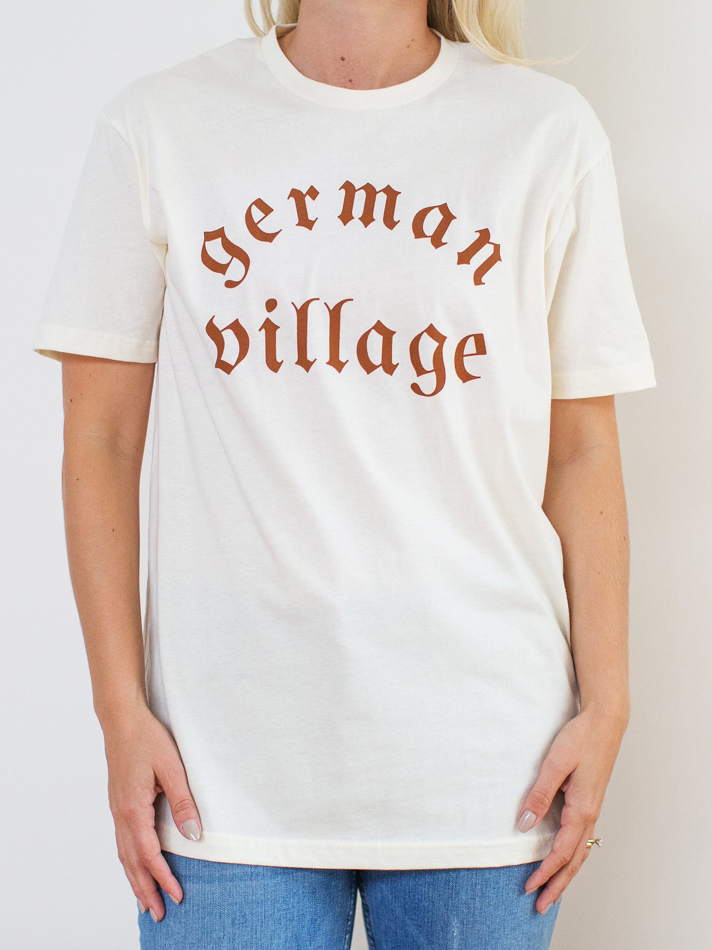 Model wearing cream colored cotton crewneck tee with German Village graphic at front